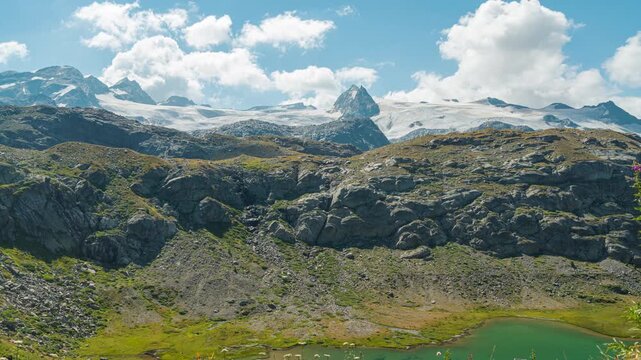 Timelapse of moving clouds over a vast glacier in Aosta Valley, Italy, on a peak named "Rutor". Mountain landscape with rocks, peaks, vegeation and lake. Summer sunny day, with blue sky.