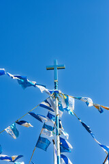 A striking blue cross, covered in glittering material, stands tall against a clear blue sky, surrounded by blue and white prayer flags.