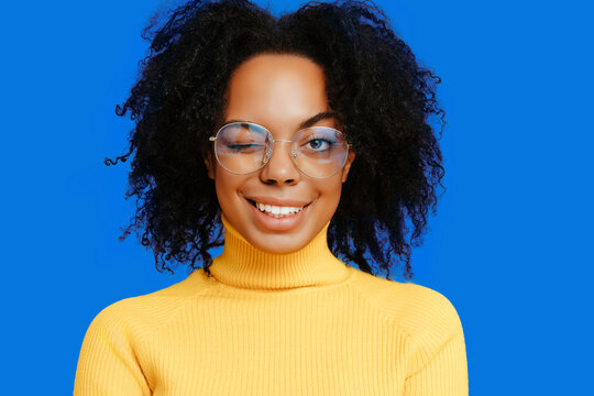 Playful African American woman in yellow turtleneck and round glasses winks with a bright smile against vivid blue background, radiating confidence and charm.
