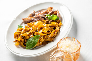 Rich, fresh tagliatelle pasta with crispy meat (possibly bacon or guanciale), raw egg yolk, and basil leaves on a white plate. Gourmet Italian meal close-up