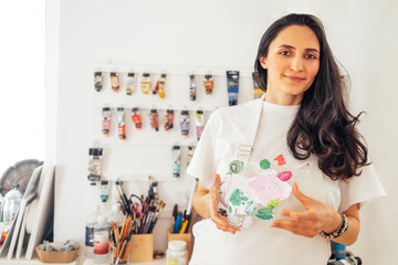 Female artist holding a painted glass vase in a bright studio, surrounded by art supplies and colorful paint tubes, showcasing creativity and artistic expression