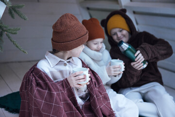 Three children wearing cozy winter clothing, sitting together indoors, enjoying warm drinks from mugs, surrounded by a festive atmosphere, conveying warmth and togetherness
