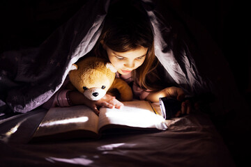 Young girl with teddy bear reading a book under a blanket with a flashlight, creating a cozy atmosphere, showcasing the joy of storytelling and imagination