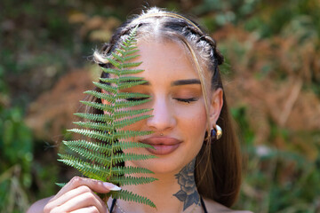 A beautiful young woman covers half her face with a green fern leaf. The woman is happy and has her...