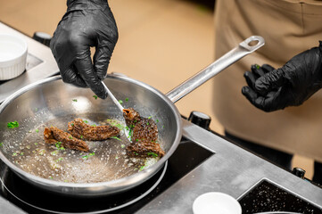 Professional chef in black gloves pan-frying gourmet morel mushrooms and herbs in a stainless steel skillet