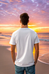 Man in white shirt stands on beach facing sunset with colorful sky and ocean in the distance view