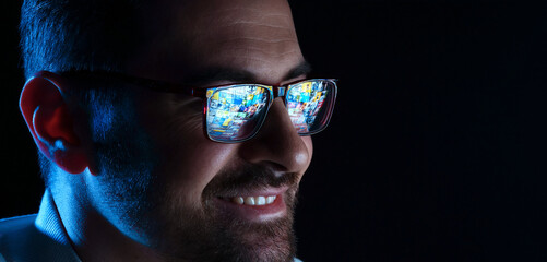 Smiling man wearing glasses with reflection on black background in color light, closeup. Space for text