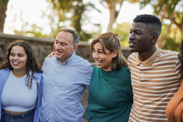 Group of multiracial people with different ages hugging each other outdoor with city park in background - Multi generational friends, social inclusion and community concept