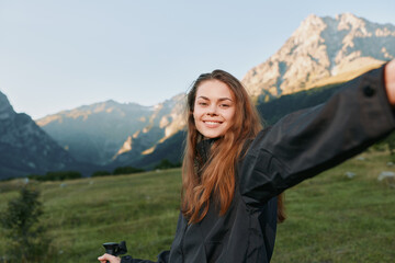 Fototapeta premium A cheerful young woman stands in a scenic mountain meadow, smiling as she takes a selfie with a compact camera, capturing a bright and energetic outdoor moment.
