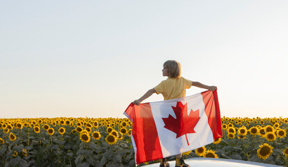 A child stands against the backdrop of a blooming sunflower field, holding a large Canadian flag...
