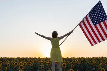 An American flag held by a woman against a backdrop of a sunset sky, sun, and a blooming field of sunflowers. The national symbol of freedom and independence of the United States of America