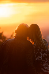 Zwei Menschen sitzen eng beieinander, R&uuml;ckenansicht, warmes Licht, romantische Stimmung, Sonnenuntergang, verschwommene Landschaft im Hintergrund.