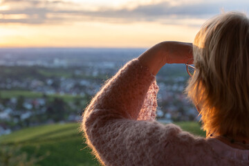 Frau mit Brille und Strickpullover blickt in den Sonnenuntergang, Landschaft im Hintergrund, warmes Licht, entspannte Stimmung.