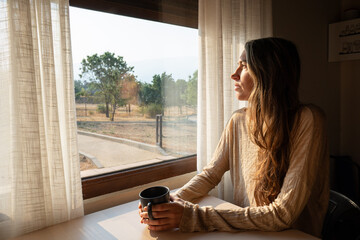 Woman drinking coffee and looking out window at sunrise in tiny house