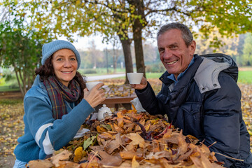 Senior couple smiling and drinking tea outdoors at a table covered with autumn leaves