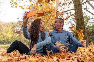 Mature couple making silly faces and taking a selfie among falling autumn leaves in a park