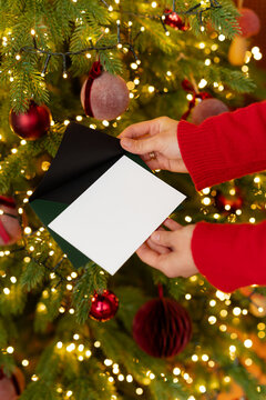 Close-up of a woman standing in front of an illuminated Christmas tree holding a blank card and envelope