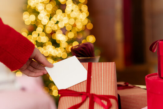 Close-up of a woman placing a blank card on a wrapped Christmas gift in front of an illuminated Christmas tree