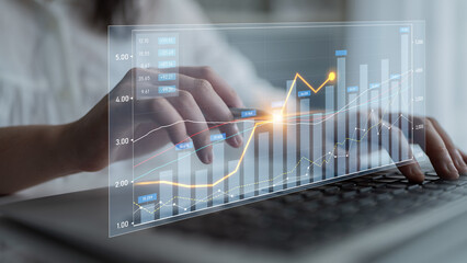 A close-up view of hands typing on a laptop with transparent digital graphs and charts showcasing...