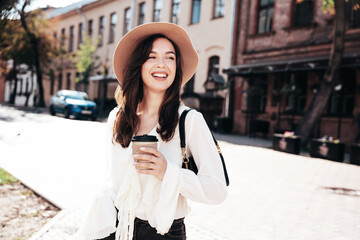 Lifestyle portrait of young stylish woman dressed in white blouse. Model walking with coffee cup in old town in sunny day. Beautiful smiling female in hat. Cheerful and happy. Takeaway coffee