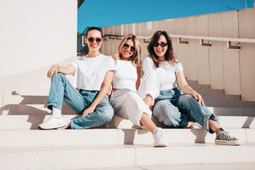 Three young beautiful smiling hipster female in trendy summer white t-shirt  and jeans clothes. Sexy carefree women posing in street. Positive models having fun, going crazy. In sunny day, sunglasses