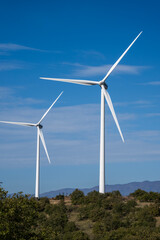 Fototapeta premium Two wind turbines stand tall on a green hillside beneath a clear sky and distant mountain range.