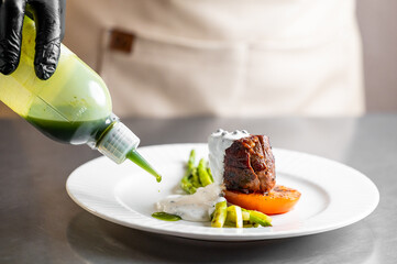 Chef in black gloves uses a squeeze bottle to drizzle green sauce on a gourmet beef tenderloin dish with asparagus and an orange element