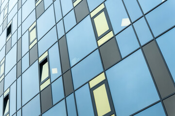 Modern building facade with blue and yellow glass windows in geometric grid pattern. Abstract urban architecture with reflective blue panels and asymmetric open windows on a contemporary glass facade.