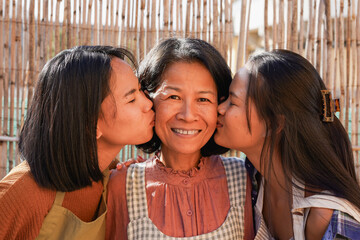 Happy asian mother having tender moment with daughters outdoor - Adult thai girls kissing mom at home patio - Family love concept