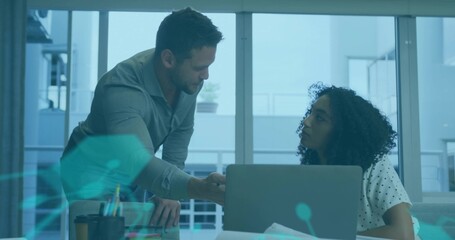 Leaning man gesturing toward woman in polka-dot blouse at office table, showing laptop, papers