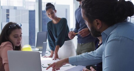 Pointing man with bun wearing blue shirt leaning over table in modern office, laptop and sketches
