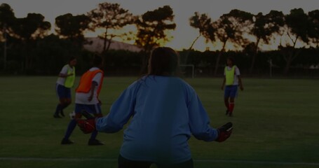 Guarding goalkeeper in blue jersey, gloves on soccer field at dusk, players in bibs dribbling ball