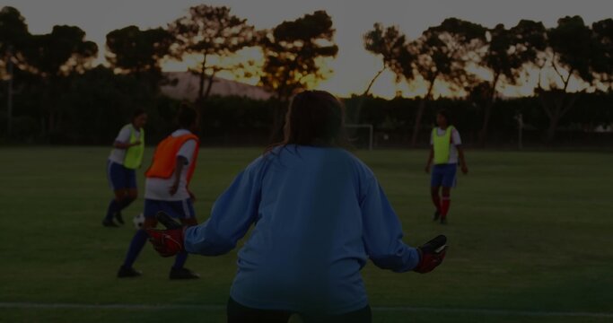 Guarding goalkeeper in blue jersey, gloves on soccer field at dusk, players in bibs dribbling ball - Powered by Adobe