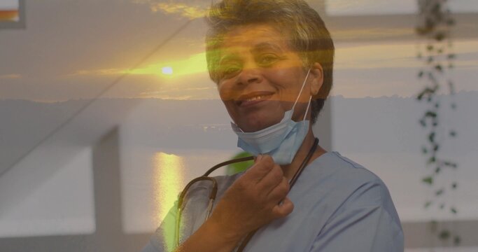 Smiling nurse wearing blue scrubs by window, showing stethoscope and lowered mask with sunset glow