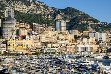Luxury yachts and modern high-rise buildings fill the glamorous Monte Carlo harbor, framed by dramatic Mediterranean mountains under clear blue skies.