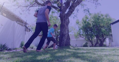 Playing father and son wearing grey shirt and blue shirt, barefoot, kicking soccer ball in backyard
