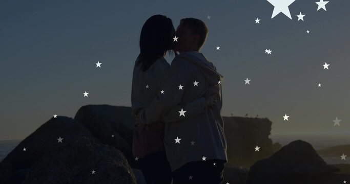 Kissing couple embracing on rocky shore at dusk, in hoodies, boulders, cliff, horizon, stars - Powered by Adobe
