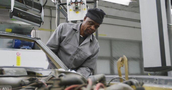 Leaning mechanic in grey coveralls and dark cap checking engine at workshop, with open hood, hoist - Powered by Adobe