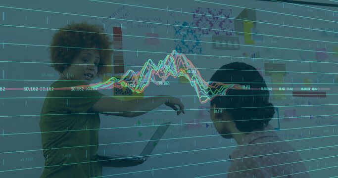 Pointing two women working at office wall with open laptop sticky notes, green top, printed blouse