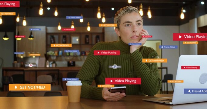 Holding phone, woman in green sweater sitting at cafe table, viewing laptop and floating alerts