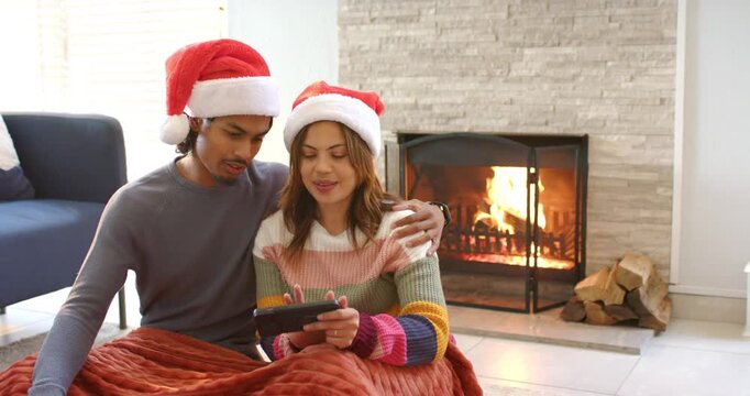 Diverse couple wearing Santa hats scrolling tablet for gift ideas, waving farewell by fireplace