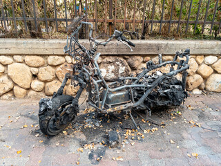 Burnt motorcycle frame after a fire on a city street. Charred metal, melted parts, and signs of destruction near a stone wall and iron fence.