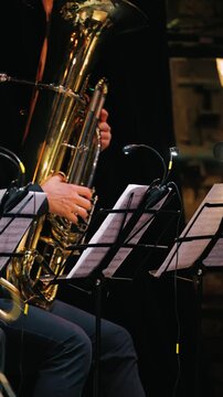 A close-up of musicians performing with a tuba and music stand. The hands of a musician playing the instrument during an orchestral performance