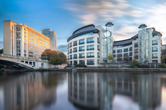 Waterfront view of modern office buildings, including a distinctive curved structure with external glass spiral staircases, reflected in the smooth, long-exposure water. - Powered by Adobe