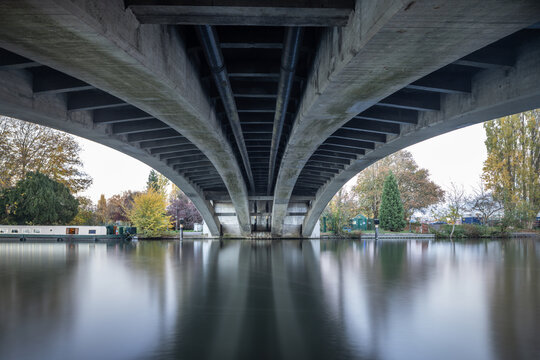 A long exposure shot looking up beneath a wide, arched concrete bridge over still water, reflecting the structure and the autumnal trees on the banks.