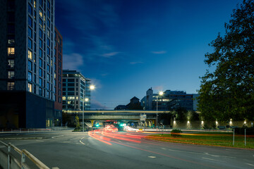 A twilight scene of a city intersection with modern buildings on the left, looking toward a bridge and underpass with blurred light trails of cars.