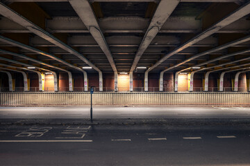 A symmetric, low-angle view of an underpass or tunnel, featuring exposed structural beams, brick walls, and a road surface with a single lamp post.