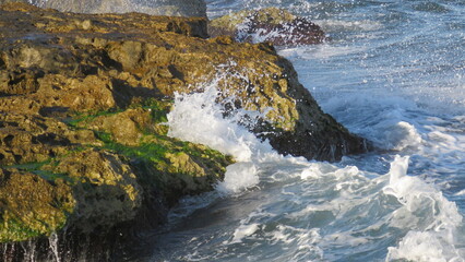 Olas rompiendo con fuerza contra rocas costeras de color ocre y musgo verde, creando salpicaduras de agua y espuma bajo el sol intenso.