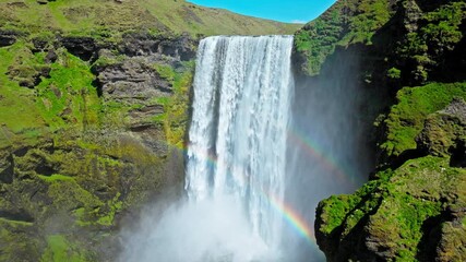 Aerial view of Skogafoss waterfall with rainbow forming in the mist. Drone footage capturing powerful white water plunging into the canyon below steep mossy cliffs. - Powered by Adobe