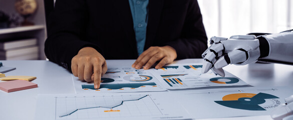 A business professional collaborates with a robotic hand while analyzing various charts and graphs, showcasing the intersection of technology and modern work in an office environment.Noogenesis